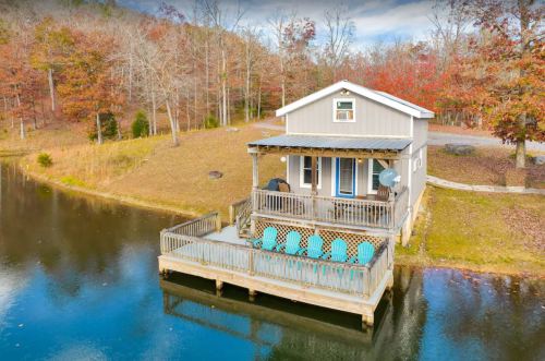 A cozy cabin on a lake with a deck and chairs, surrounded by autumn trees and a serene landscape.