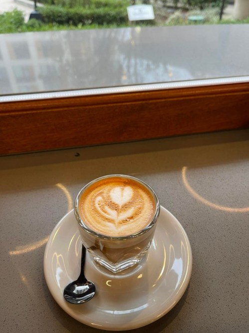 A close-up of a latte in a glass cup with a heart design, sitting on a white saucer with a spoon beside it.