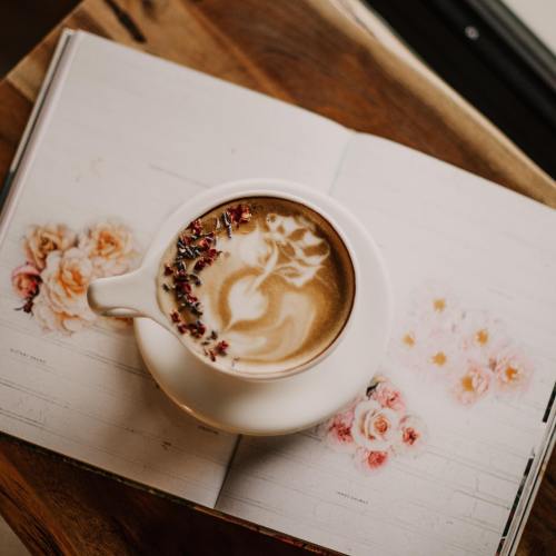 A latte with floral art sits on an open book featuring rose illustrations.