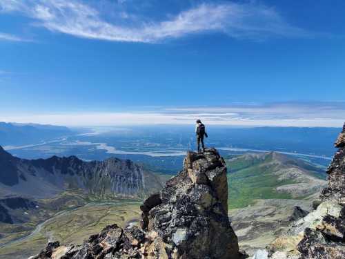 A hiker stands on a rocky peak, overlooking a vast landscape of mountains and a river under a clear blue sky.