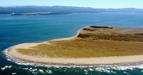 Aerial view of a sandy peninsula extending into a blue ocean, surrounded by gentle waves and distant hills.