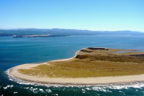 Aerial view of a sandy peninsula extending into a blue ocean, surrounded by gentle waves and distant mountains.