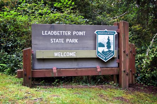 Sign for Leadbetter Point State Park, featuring "WELCOME" and a state emblem, surrounded by greenery.