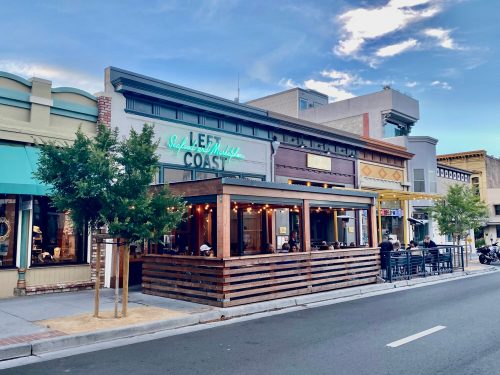 A restaurant with outdoor seating and a sign reading "Left Coast" on a street with shops and blue sky.