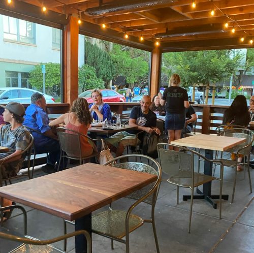 A lively outdoor restaurant scene with diners at wooden tables, string lights overhead, and a server attending to guests.