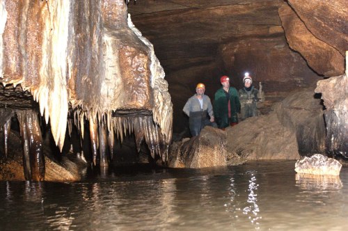 Three people in helmets stand in a cave with stalactites and a water pool, surrounded by rocky formations.