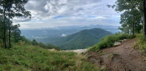 A panoramic view of rolling green mountains under a cloudy sky, with a foreground of grass and rocky terrain.
