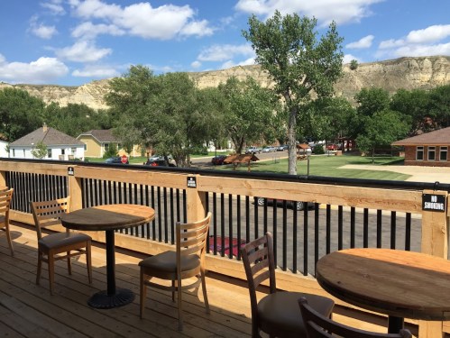 A scenic view from a wooden deck with empty tables, overlooking trees and a hillside under a blue sky with clouds.