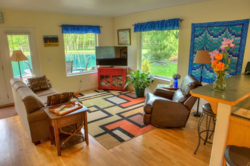 Cozy living room with a sofa, armchair, colorful rug, and large windows overlooking a pool and garden.