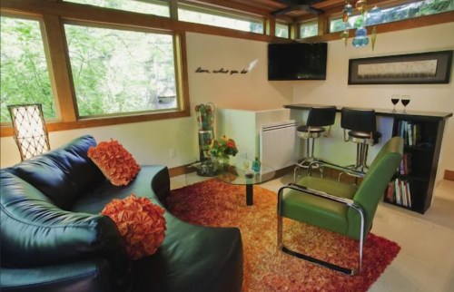 Cozy living space with a black sofa, green chair, bar area, and large windows overlooking greenery.
