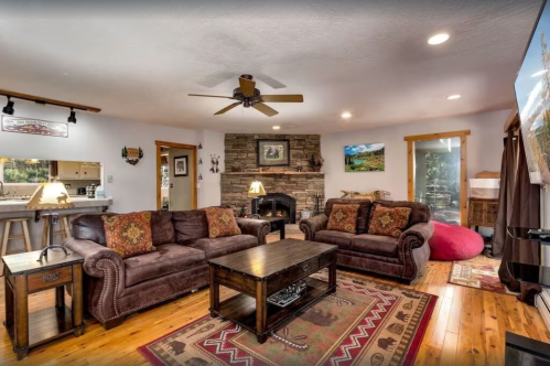 Cozy living room with brown sofas, a wooden coffee table, stone fireplace, and warm lighting. Rustic decor and wooden accents.
