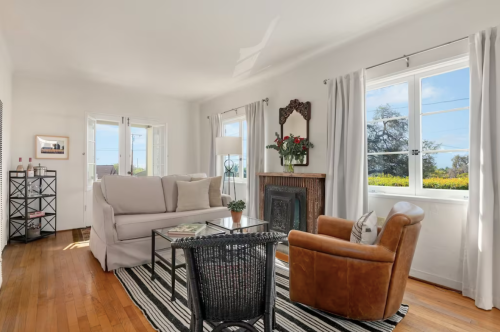 Bright living room with a beige sofa, leather chair, glass coffee table, and large windows overlooking greenery.