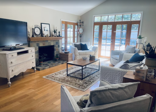 Bright living room with a TV, fireplace, and comfortable seating, featuring large windows and wooden floors.