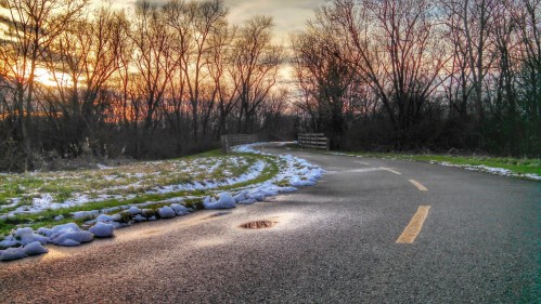 A winding path through a winter landscape, with snow patches and trees silhouetted against a colorful sunset.