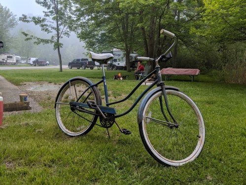 A vintage bicycle stands on green grass near a campsite with trees and campers in the background.
