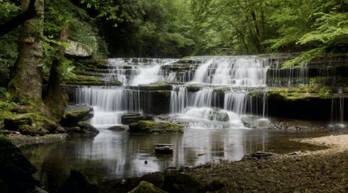 A serene waterfall cascades over rocky steps, surrounded by lush green trees and a calm riverbed.
