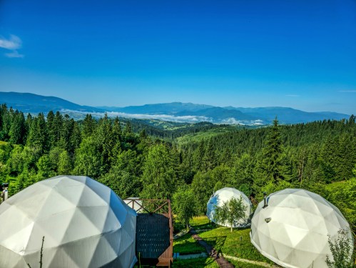 Luxury glamping dome tents on a mountain site in the morning, with a lush green forest and valley under a clear blue sky.