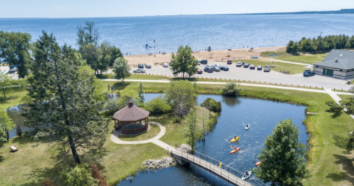 Aerial view of a beach with a gazebo, pond, and kayakers, surrounded by trees and a parking area.