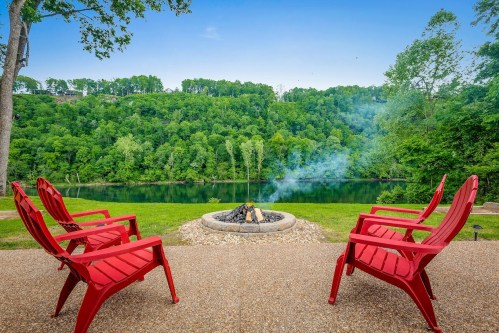 Red chairs surround a fire pit by a calm river, with lush green hills in the background and smoke rising from the fire.