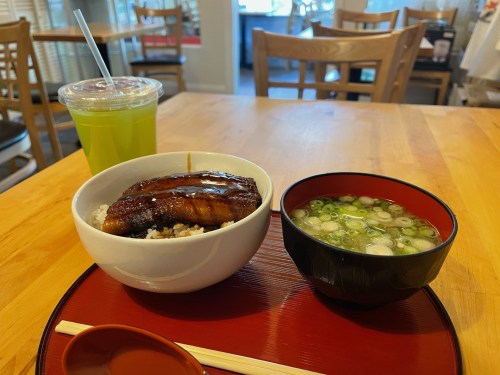 A bowl of rice topped with grilled eel, served with a side of green onion soup and a drink on a wooden table.