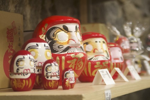 A display of colorful Daruma dolls in various sizes on a wooden shelf, with a stone wall in the background.
