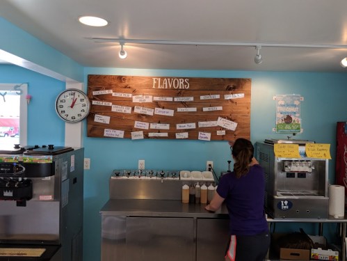 A person stands at an ice cream counter with a flavor menu displayed on a wooden board above.