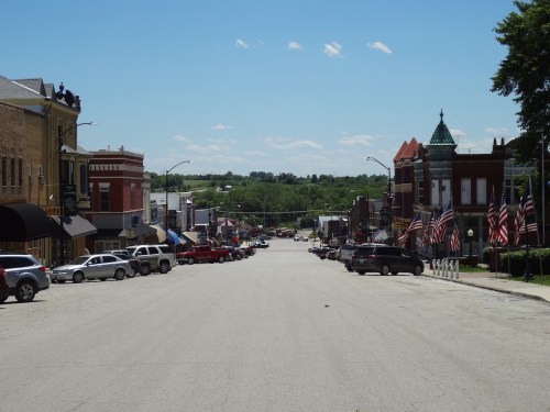 A wide view of a small town street lined with shops, parked cars, and American flags under a clear blue sky.