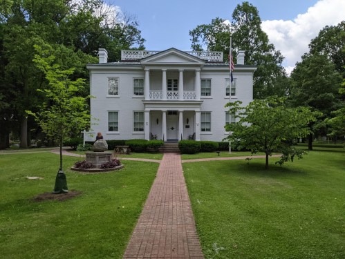 A white two-story house with a front porch, surrounded by green trees and a brick pathway leading to it.