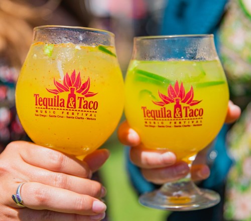 Two hands hold colorful cocktails in branded glasses at the Tequila & Taco Music Festival.