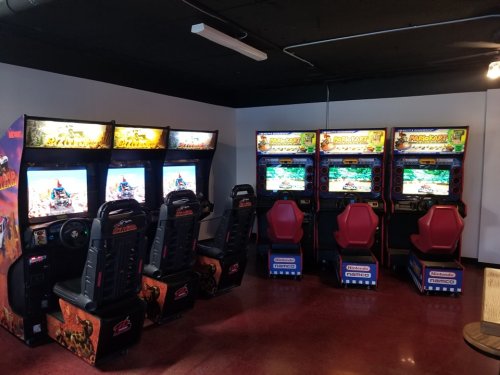 A corner of an arcade featuring multiple video game machines with racing and adventure themes.