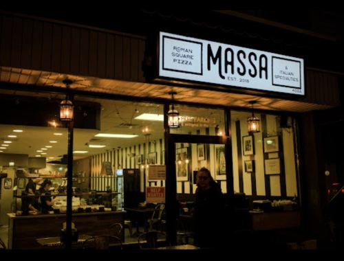 A cozy pizzeria named Massa, featuring a bright sign and inviting interior, with a person standing outside.