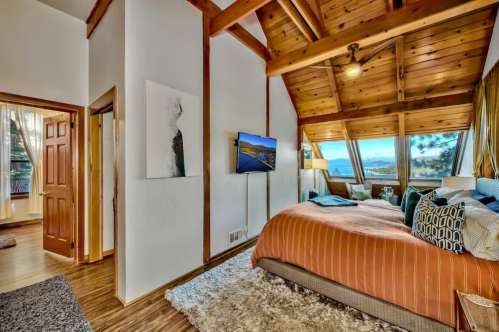 Cozy bedroom with wooden beams, large windows, a TV, and a warm color palette, overlooking a scenic view.
