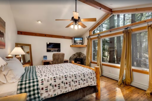Cozy bedroom with a wooden bed, large windows, and a stone fireplace, surrounded by trees.
