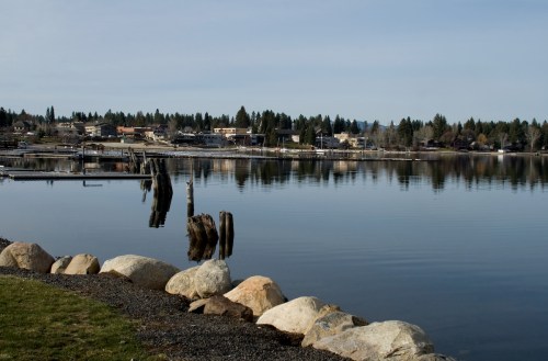 A calm lake reflecting a small town and trees, with rocks in the foreground and docks in the background.