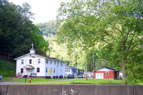 A white church with a cross, surrounded by green trees and a few buildings, set against a mountainous backdrop.