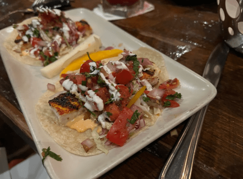 Two tacos on a plate, topped with grilled fish, fresh salsa, lettuce, and drizzled with sauce.