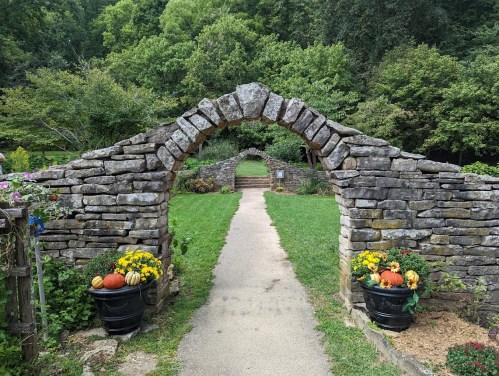 A stone archway leads to a grassy path, flanked by flower pots with pumpkins and flowers, surrounded by lush greenery.