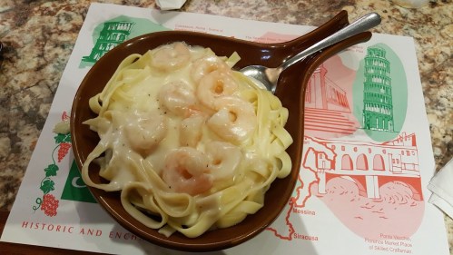 A bowl of fettuccine pasta topped with shrimp in a creamy sauce, served on a colorful placemat.