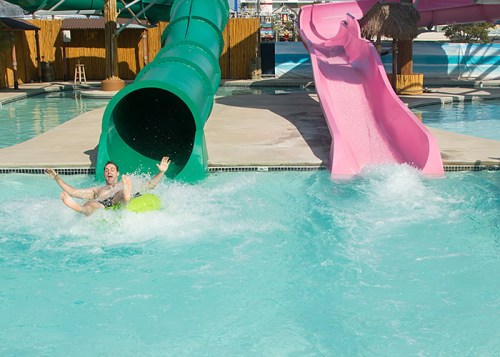 A person slides down a water slide into a pool, splashing water around them. Two colorful slides are visible.