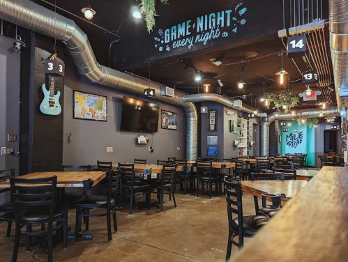 Interior of a cozy restaurant with wooden tables, black chairs, and a game night theme, featuring wall decor and a TV.