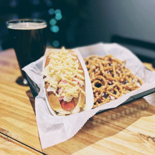 A hot dog topped with sauerkraut and fries, served with a side of pretzels and a glass of dark beer.