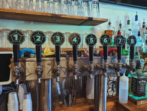 A row of eight beer taps with numbered handles, set against a backdrop of bottles and glasses on a bar.