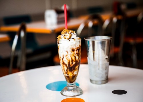 A tall glass of ice cream sundae topped with whipped cream and chocolate sauce, next to a metal milkshake cup.