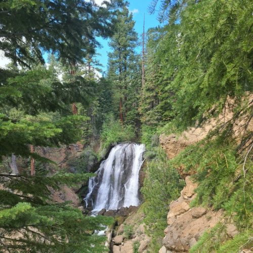 A serene waterfall cascades down rocky cliffs, surrounded by lush green trees and a clear blue sky.