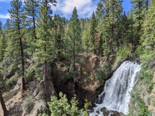 A serene waterfall cascades down rocky terrain, surrounded by lush green trees and a clear blue sky.