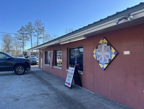A storefront with a colorful quilt pattern sign, parked cars, and a sign directing to a business entrance.