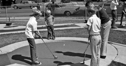Black and white photo of children playing mini-golf, focused on one boy preparing to putt while others watch.