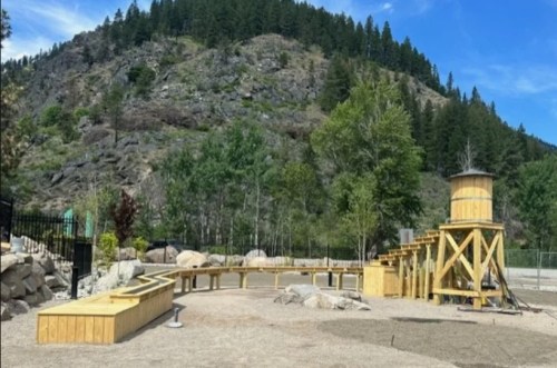 A wooden playground structure with a water tower, surrounded by trees and mountains under a clear blue sky.