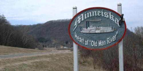 Sign for Minneiska, "Head of Old Main River," with a scenic backdrop of hills and a road in the foreground.