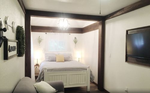 Cozy bedroom with a white bed, soft lighting, and wooden accents, featuring a TV and decorative plants.